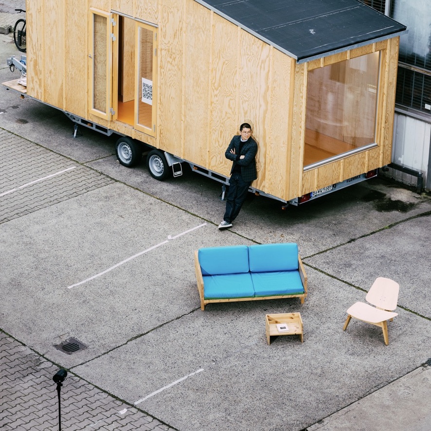 Der Berliner Architekt und Van Bo Le-Mentzel lehnt an dem von ihm entworfenen Tiny Haus. Vor ihm auf dem Asphalt: Luxusmöbel zum Selberbauen aus Holz aus dem Baumarkt, die er ebenfalls entworfen hat.
