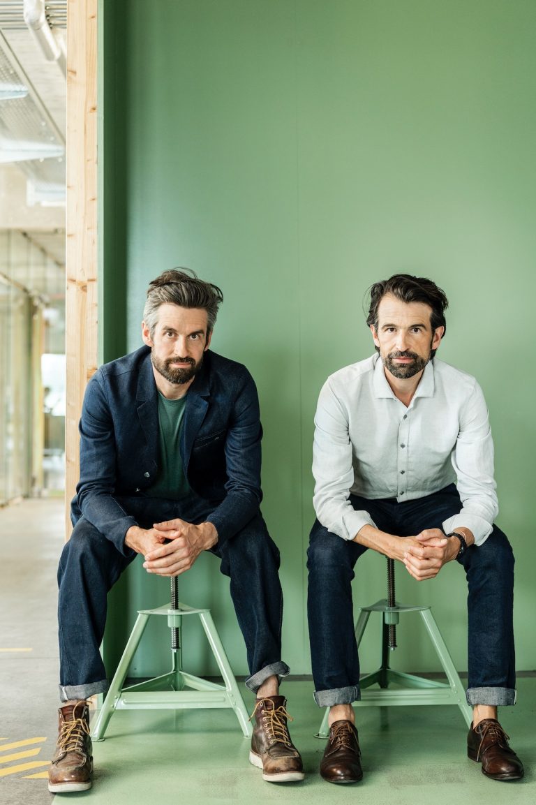 Die Brüder Daniel Freitag (li.) und Markus Freitag (re.) gründeten 1993 ihr Designunternehmen FREITAG in Zürich. Hier sitzen sie nebeneinander auf grünen Hockern vor einer grünen Wand.